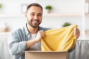 Happy Caucasian guy unboxing cardboard parcel, satisfied with successful shopping, taking out new sweater, receiving new purchase at home. Online store and delivery concept photo