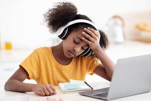 Upset black school girl doing homework, sitting at kitchen table in front of laptop, using wireless headset, looking at notepad and touching her head, burnout at school concept, copy space photo