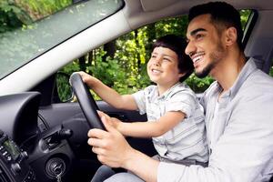 Cute Little Boy And His Young Arab Father Riding Car Together, Little Male Child Sitting On Dad's Lap In Automobile, Caring Middle Eastern Daddy Teaching Happy Kid How To Drive, Side View Shot photo