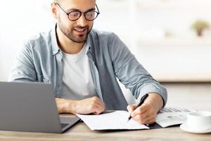 Young Caucasian businessman taking notes while working on laptop computer at home office. Freelancer noting down information from internet, planning working schedule, sitting at desk indoors photo