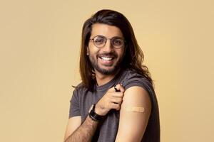 Happy long-haired arab guy in casual got vaccinated against coronavirus at hospital or clinic, sitting on chair over beige studio background, showing his shoulder with adhesive band on, copy space photo