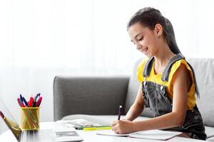 Portrait of smiling schoolgirl sitting on couch at desk with pc, writing essay or drawing picture in paper notebook. Pupil studying or taking notes in her diary, making checklist, free copy space photo