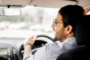 Back view of middle-eastern long-haired guy driving auto, looking at copy space and smiling, shot from automobile back seat, closeup. Cheerful arab man enjoying his brand new car photo