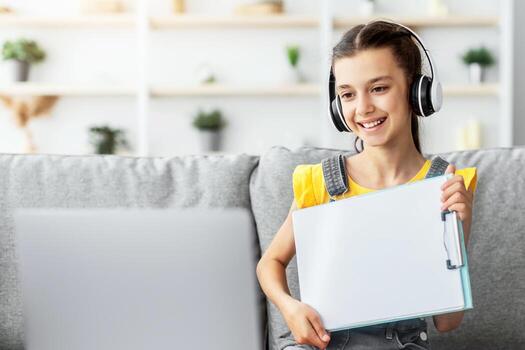 Smiling girl having call with teacher, holding flipchart with paper showing done task result using laptop. Child studying remotely. Creativities lessons, homeschool photo