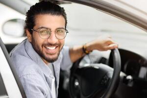 Car leasing, buying, retail, renting concept. Portrait of happy arab guy in casual outfit getting into brand new auto, placing hand on wheel, cheerfully smiling at camera, side view, copy space photo