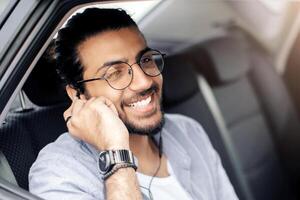 Cheerful arab guy sitting at car back seat, listening to music while going somewhere by transportation service, using earphones and mobile application on smartphone, enjoying view, copy space photo