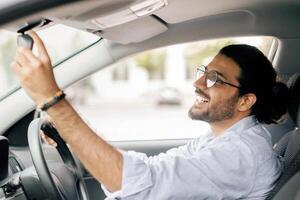 Positive middle-eastern long-haired guy driver adjusting rear view mirror in his brand new car, side view. Happy arab young man looking at back view mirror while driving modern comfortable auto photo
