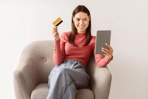Online shopping, remote banking concept. Cheerful young lady with credit card and tablet pc sitting in armchair, making purchases on internet, using web store application, making contactless payment photo