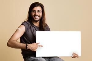 Handsome middle-eastern young man with long hair and glasses got vaccinated against COVID-19, holding horizontal white blank board, sitting on chair over beige studio background, copy space, mockup photo