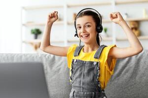 Portrait of smiling child wearing headset showing yes gesture playing online game or studying, shaking and raising clenched fists up in the air, looking at camera, using computer free copy space photo