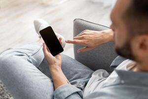 Unrecognizable young guy using smartphone with empty screen at home, mockup for website or mobile app design. Closeup of millennial man holding modern gadget, space for advertisement photo