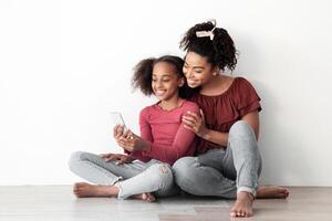 Beautiful black mother and daughter using mobile phone together, sitting on floor, hugging and taking selfie for social media, enjoying time together at home, white empty wall backgound, copy space photo