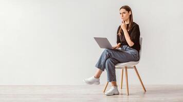 Troubled Caucasian woman sitting on chair with laptop computer, making mistake in online project, feeling stressed, thinking over solution to problem against white wall, copy space. Banner design photo