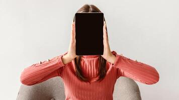 Young woman holding tablet pc with blank screen in front of her face on white studio background, mockup for app or website. Millennial lady presenting space for advertisement on touch pad photo