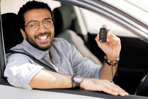 Cheerful middle-eastern long-haired guy with glasses sitting inside car and showing automatic key, positive arab man renting automobile for travelling, having car ride, closeup portrait photo