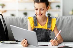 Portrait of smiling junior school kid girl holding digital tablet studying online during virtual class learning at home, doing homework and taking notes in her copybook or having call with tutor photo