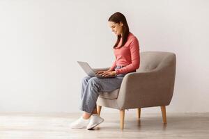 Happy young lady sitting in armchair, using laptop, studying remotely or having business meeting, free space. Millennial Caucasian woman having online class or watching webinar photo