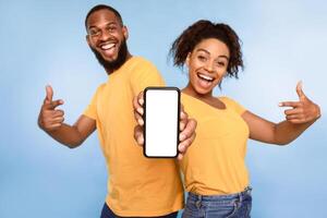 Mobile application ad. Excited African American couple pointing at cellphone with blank screen, recommending new app on blue studio background, selective focus. Mockup for website photo