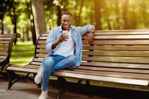 Smiling black man sitting on bench at park and using his smartphone, copy space photo