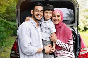 Happy Muslim Family With Little Son Posing Outdoors Near Car With Open Trunk, Cheerful Arab Father, Child And Mother In Hijab Embracing And Smiling At Camera, Enjoying Automobile Trips Together photo