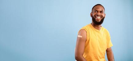Cheerful vaccinated African American man with plaster bandage on his shoulder after flu vaccine injection on blue studio background, banner design with free space. Flu immunization campaign photo
