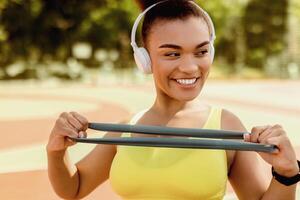 Outdoors Training Concept. Smiling black lady holding latex loop doing exercises for arms and chest muscles. Portrait of young fit woman working out with resistance band, wearing wireless headset photo