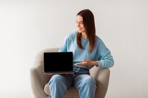 Smiling millennial woman in casual clothes showing laptop with empty screen, sitting in armchair on white studio background, mockup for website. Young lady presenting portable computer with template photo