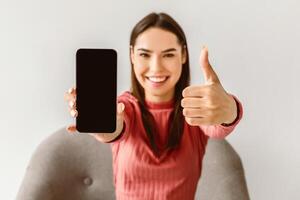 Happy Caucasian lady showing cellphone with empty screen and gesturing thumb up, recommending new mobile app or website on white studio background, mockup for advertisement photo
