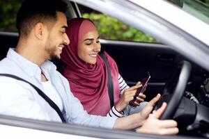 Young Muslim Spouses Riding Car And Using Smartphone Together, Happy Middle Eastern Couple Browsing Online Maps Or Gps Navigation On Mobile Phone, Planning Travel Route, Through Window Shot photo
