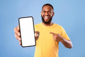 Handsome happy African American guy pointing at smartphone with blank screen on blue studio background, mockup for website or new mobile app. Cellphone display template. Space for advertisement photo