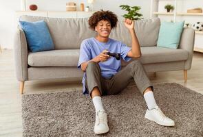Full length of cheery black adolescent enjoying videogame on playstation, having fun at home. Positive Afro teenager with joystick sitting on floor, playing computer arcade. Lockdown hobbies concept photo