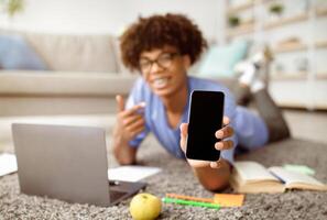 Positive black teenager lying on floor with laptop, pointing at smartphone with blank screen at home, mockup for mobile app or website design. Remote studies during coronavirus lockdown concept photo