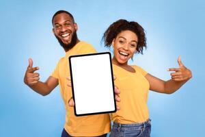 Joyful African American couple pointing at tablet screen with mockup for design, advertising new app or website over blue studio background. Touch pad display template, space for your ad photo