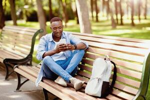 Smiling African American guy with smartphone and backpack resting on bench at park, blank space. Funky black man in casual outfit using mobile device for online communication outside photo