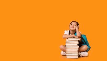 Chinese Elementary School Girl Sitting At Books Stack Posing On Yellow Background. Studio Shot, Free Space For Text photo