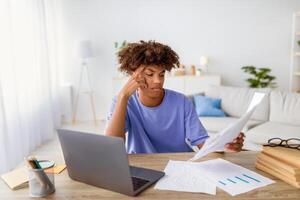 Bored irritated black teenager sitting at table with papers, using laptop to write coursework, studying online, making hard home assigment, preparing for exam. Remote schooling concept photo