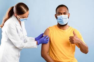 African American guy in face mask showing thumb up gesture after flu vaccine shot, recommending anti flu vaccination, lady doctor putting on band aid over his arm, blue studio background photo