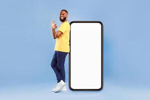Optimistic African American man showing ok gesture, leaning on huge phone with blank white screen, recommending new mobile application on blue studio background, mockup for website ad design photo