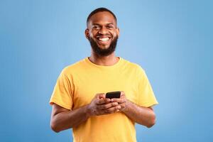 Happy young African American man using smartphone, writing message, chatting with friend, surfing internet, watching movie online, having web lesson or meeting on blue studio background photo