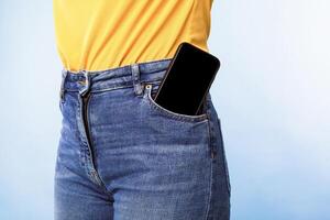 Unrecognizable woman having smartphone with empty screen in her jeans pocket on blue studio background, mockup for mobile app or website design. Space for your ad template photo