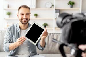 Positive male vlogger recording review of tablet computer at home, mockup. Happy blogger showing electronic device with empty screen at camera, promoting new model of gadget photo
