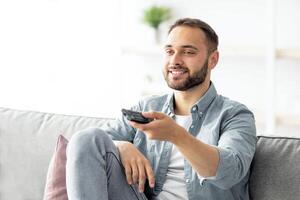 Weekend leisure activities. Millennial Caucasian man with remote control switching channels on TV at home. Happy young guy watching interesting program on television indoors photo