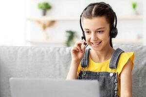 Kids And Technology Concept. Portrait of smiling girl sitting on couch at table and using laptop computer, wearing and touching headset talking in microphone having call or playing online game photo