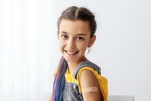 Successful Covid-19 Vaccination Concept. Vaccinated Smiling Teenage Girl Showing Arm With Adhesive Bandage After Injection, Posing Sitting At Clinic Hospital, Blurred Background, Free Copy Space photo