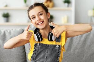 Smiling girl in headphones enjoying elearning sitting on sofa at table showing thumbs up recommending e-study and online school for children, using modern tech. Clever kid and self-education concept photo