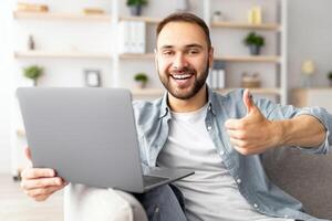Excited millennial guy using laptop at home, showing thumb up gesture, recommending online work or studies. Young male freelancer communicating on web, taking part in business meeting photo