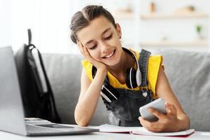 Modern Teen's Lifestyle. Portrait of Smiling Digital Native Girl Texting On Phone Doing Homework Sitting At Desk Using Laptop At Home. Cheerful Student Resting On Sofa Scrolling Social Media News Feed photo