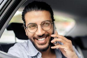 Closeup of cheerful young indian man sitting in taxi back seat, talking on cellphone with lover and smiling, shot from window, copy space. Handsome long-haired arab guy using transportation service photo