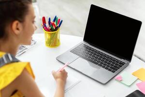 Over the shoulder portrait of happy smiling student girl sitting at desk, doing homework, writing in her notebook and looking at black blank laptop screen, studying remotely, selective focus, mock up photo