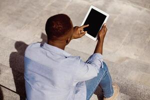 Young black guy using tablet computer with empty screen on stone stairs outdoors, mockup for website design. Millennial African American man pointing at touch pad with space for your template photo